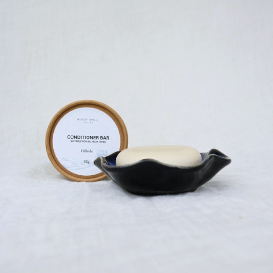Bar of soap in a black dish with a container labeled 'Conditioner Bar' on a white background